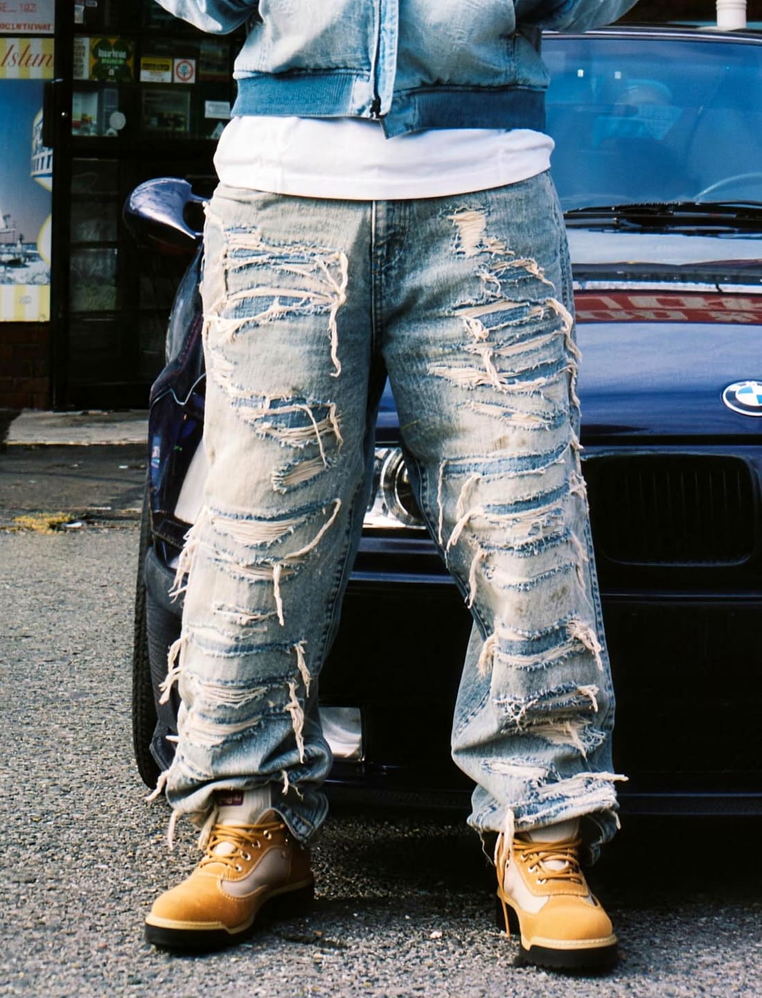 A person wearing heavily distressed light-wash blue jeans with ripped details and frayed edges, paired with tan boots, standing in front of a dark blue car.