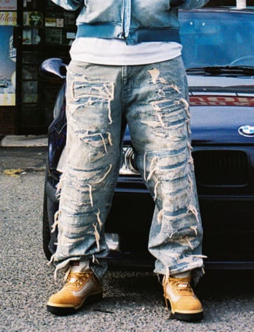 A person wearing heavily distressed light-wash blue jeans with ripped details and frayed edges, paired with tan boots, standing in front of a dark blue car.